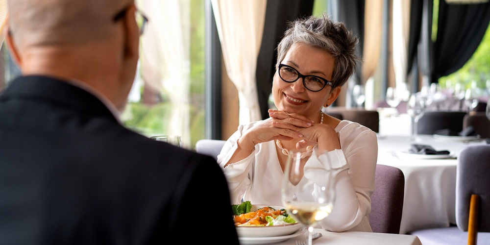 A woman smiling warmly at a dining companion in a bright, elegantly set restaurant, with a plate of food and a glass of white wine on the table.