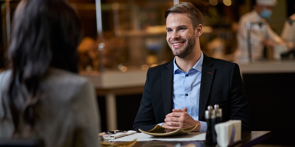 A guest smiling and engaging with a another guest in a full-service dining room, menu on the table and kitchen visible in the background.