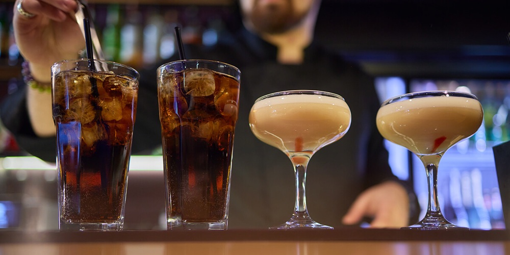 Four drinks lined up on a bar counter — two iced soft drinks and two coupe glasses with a creamy cocktail — prepared by a bartender.