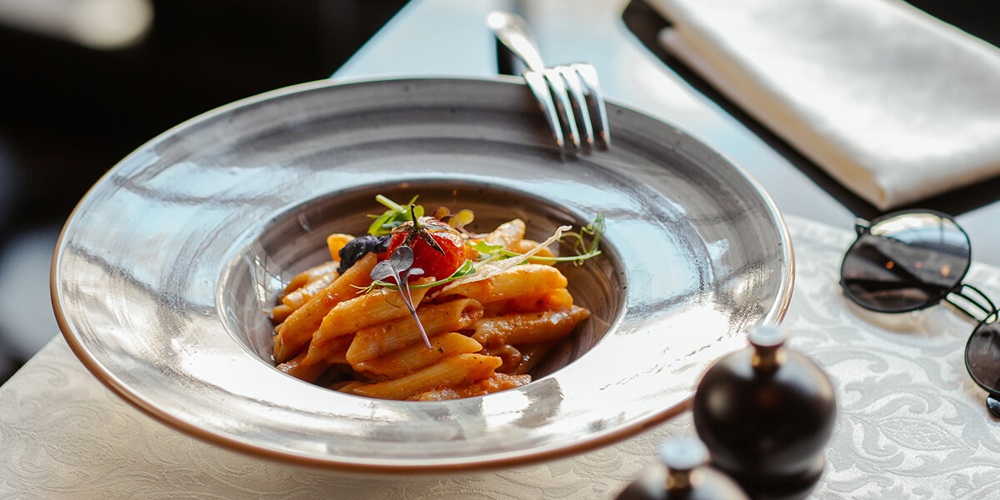 Penne pasta in tomato sauce served in a wide-rim metallic bowl, garnished with cherry tomato and microgreens, at a fine-dining restaurant table.