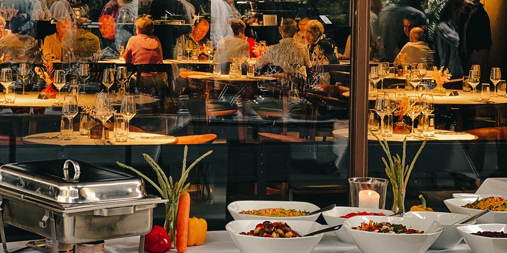 A buffet setup in the foreground with chafing dishes, fresh vegetables, and white bowls of prepared dishes, with a full dining room visible through the glass behind.