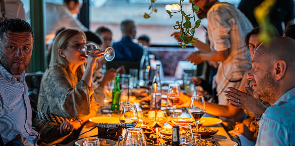 Guests sharing a lively dinner at a long table under warm amber lighting, with multiple dishes, wine, and a waiter attending to the group.